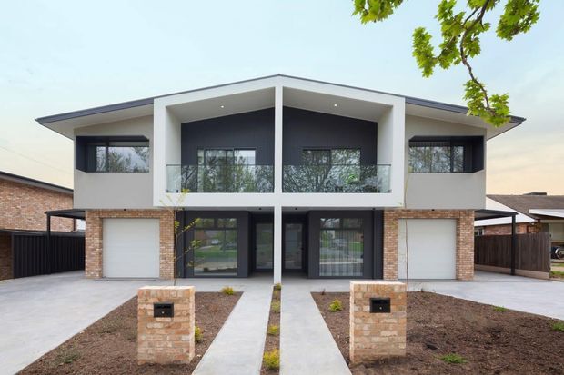 Front exterior view of a contemporary two-storey suburban residence with a broad low-pitched roof, strong white rectangular framing, dark charcoal wall cladding, extensive glazing, and warm brickwork at the lower level. The frontage includes a central balcony or recessed upper terrace, double garage access, paved paths, and low brick letterbox pillars.