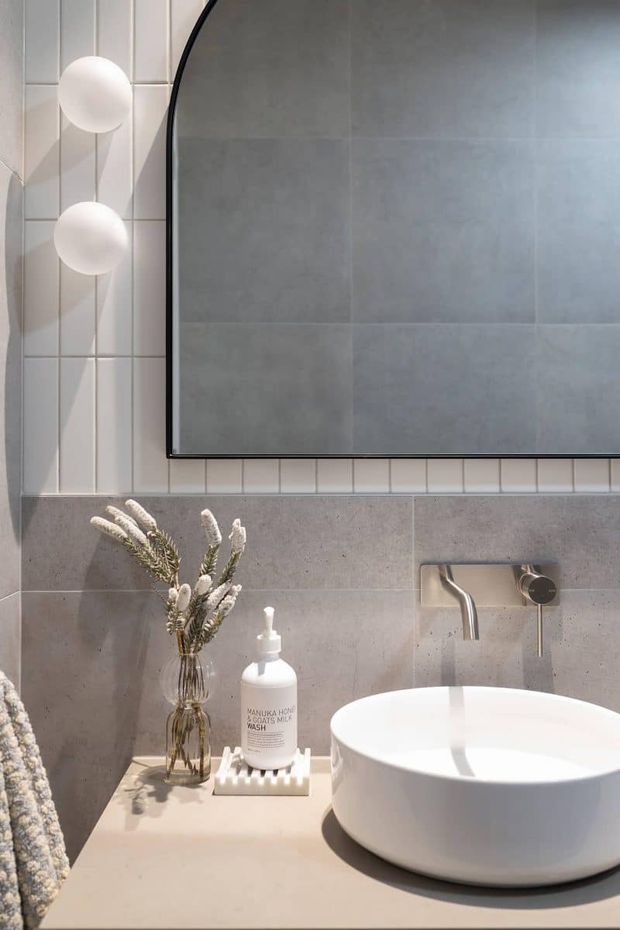 Close-up view of a refined contemporary bathroom vanity. A white vessel basin sits on a pale stone benchtop with a wall-mounted chrome tap, against grey and white tiled walls. A large black-framed arched mirror and spherical wall lights add strong graphic details, while dried botanical stems provide a subtle natural accent.
