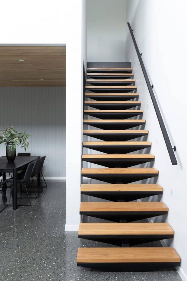 A contemporary open-riser staircase with warm timber treads and dark sculptural supports rises beside a white wall with a slim dark handrail. An adjacent dining area is visible, featuring a dark table, upholstered chairs, grey vertically panelled walls, and a terrazzo-style floor.