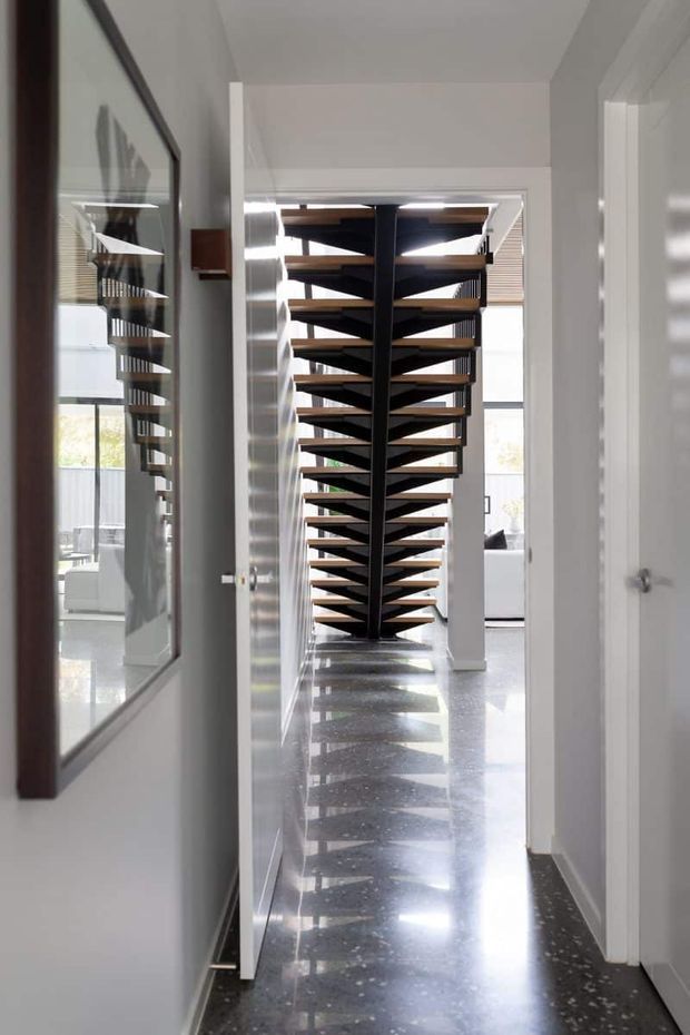 A bright contemporary hallway with a dramatic open-riser, sculptural timber-and-dark-metal staircase at the far end. White walls and doorways frame the corridor, while a polished dark terrazzo floor reflects the staircase. A large framed artwork or mirror is mounted along the left wall, and glimpses of a light-filled living area and exterior balcony are visible beyond the stairs.