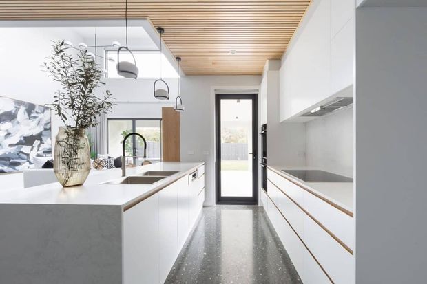Bright contemporary kitchen with a long central island, white flat-panel cabinetry, integrated appliances, a stone island benchtop, and a dark polished terrazzo-like floor. A timber slatted ceiling adds warmth, while black-framed glazing and pendant lights provide contrast. The kitchen opens toward a rear glazed door and adjoining living area.