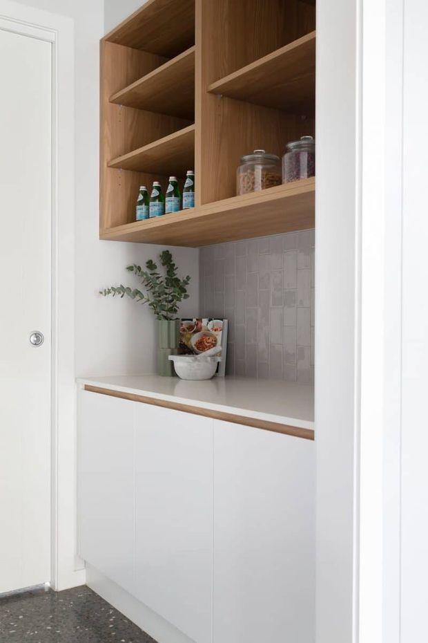 A compact pantry or kitchen storage nook viewed through a doorway, featuring white lower cabinetry, open natural timber shelving, a light benchtop, and a pale grey rectangular tiled splashback. Jars, bottles, and decorative greenery are arranged on the shelves and counter. The space is bright and clean with a restrained, contemporary look.