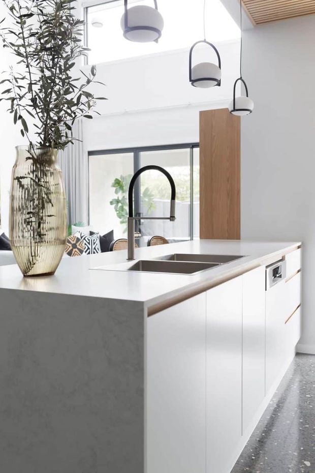 Bright contemporary kitchen centered on a large island with a white stone benchtop, integrated double sink, matte white cabinetry, and a black gooseneck tap. A warm timber vertical cabinet, dark-framed glazing, sculptural pendant lights, and a slatted timber ceiling detail add contrast. A tall decorative indoor plant and vase sit on the island.