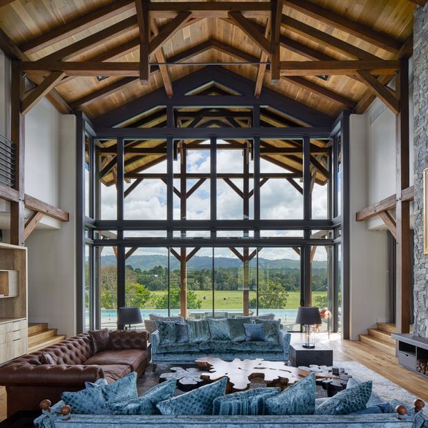 A dramatic double-height living room with exposed timber roof trusses and beams, extensive black-framed glazing, a stone feature wall, and wide views across a green rural landscape. The room is furnished with a large blue sectional sofa, a brown leather sofa, a cowhide rug, and dark occasional tables.