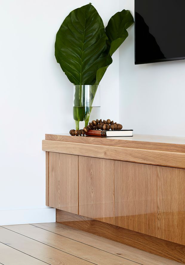 Close-up interior view of a minimalist timber sideboard or media console with vertical wood-grain cabinet doors and a thick timber top. A clear glass vase holding large green leaves sits on top alongside stacked books and decorative objects. The console is positioned against a white wall with part of a wall-mounted black television visible above it. Light timber flooring and a recessed plinth complete the composition.