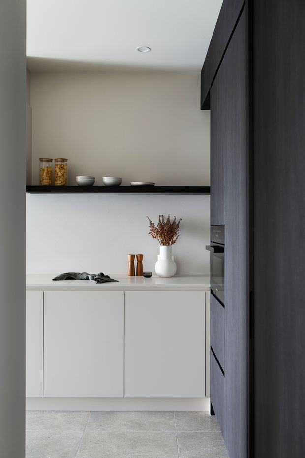 A compact, minimalist kitchen with flat-panel pale cabinetry, a light stone or composite benchtop, dark vertical timber cabinetry on the right, and a black open display shelf. Integrated appliances and simple neutral styling create a clean, refined look. Large-format grey floor tiles and recessed ceiling lighting complete the space.