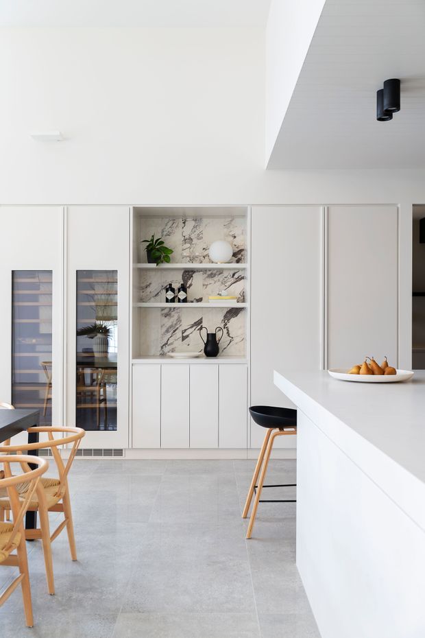 Bright, minimalist contemporary kitchen with a large white island, pale cabinetry, integrated glass-front storage, and a marble-look feature recess displaying decor. The room has a light grey tiled floor, white walls and ceiling, black pendant and track-style lighting, and timber-framed dining and bar stools.