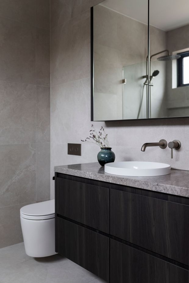 A compact contemporary bathroom with large light grey stone-look wall and floor tiles, a dark timber vanity with grey stone benchtop, white vessel basin, wall-mounted brushed metal tapware, frameless shower glazing, toilet, and a large black-framed mirrored cabinet. A small dark ceramic vase adds a restrained decorative touch.