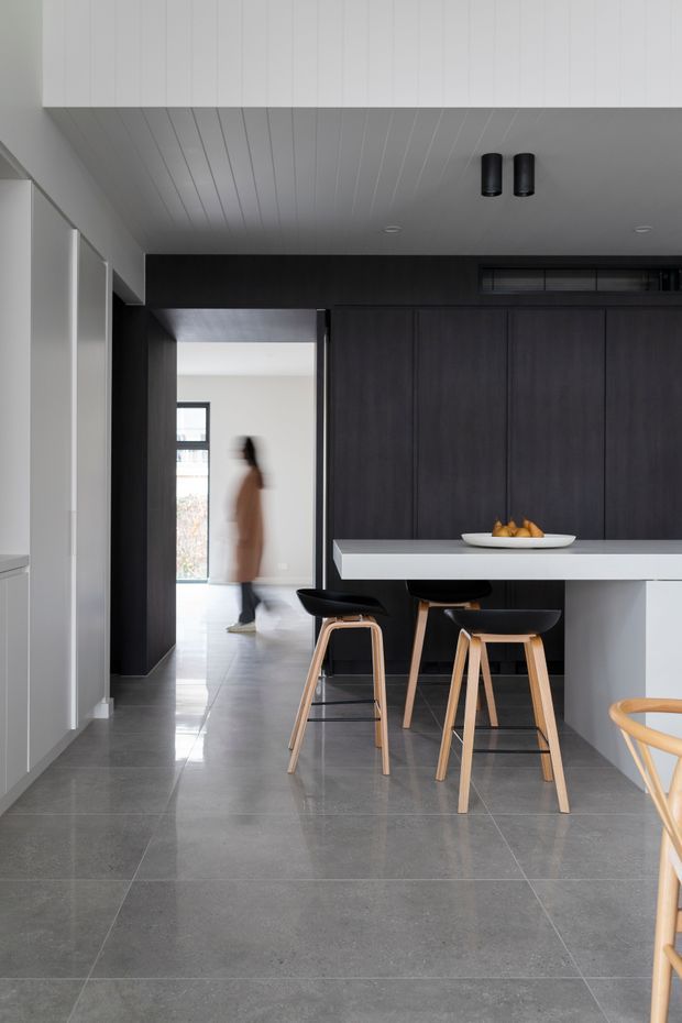 A refined contemporary kitchen with dark vertical timber cabinetry, a white island bench, black and timber bar stools, large grey floor tiles, and a white panelled ceiling. The kitchen opens to a bright hallway beyond, with a blurred person in the background. The palette is monochromatic and sophisticated with warm timber accents.