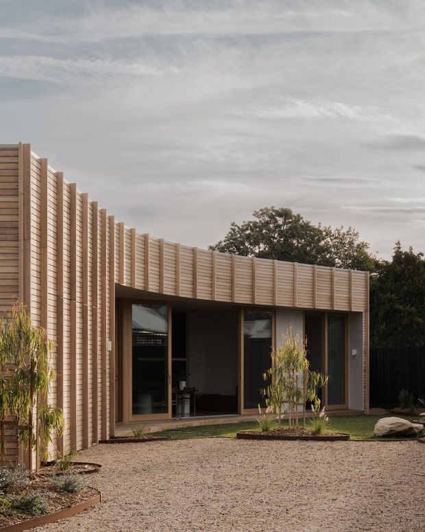 A low-rise contemporary courtyard building with a curved or gently faceted enclosure clad in repeated vertical timber boards. Large dark-framed glazed openings form a sheltered façade beneath the timber screen, opening onto a gravel courtyard with sparse landscaped planting and mature trees beyond. The restrained natural palette and sculptural timber geometry create a calm, modern exterior.