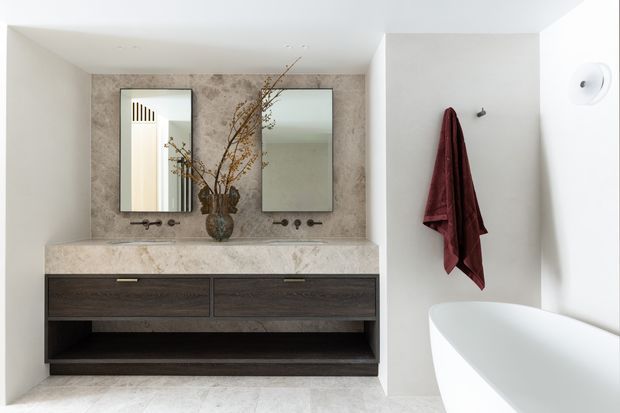 Contemporary bathroom featuring a long double vanity with dark wood drawers and open shelving, a pale stone benchtop and matching stone feature wall, twin rectangular mirrors, wall-mounted taps, and a freestanding white bathtub. The space has a restrained neutral palette with a burgundy towel providing contrast.
