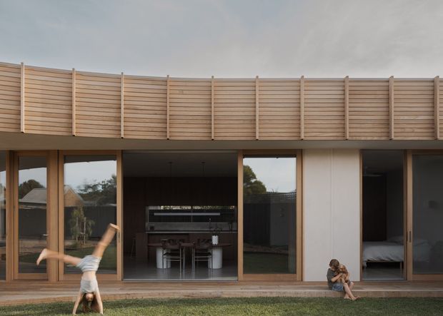 A contemporary single-storey house facade with broad floor-to-ceiling glazing opening onto a lawn. The upper level is screened with horizontal timber slats, while the lower facade combines warm timber-framed glazing, pale rendered wall panels, and dark interior finishes. A dining area and kitchen are visible through the central glazing, with a bedroom visible at the right. The image has a restrained, modern architectural character with strong indoor-outdoor connection.