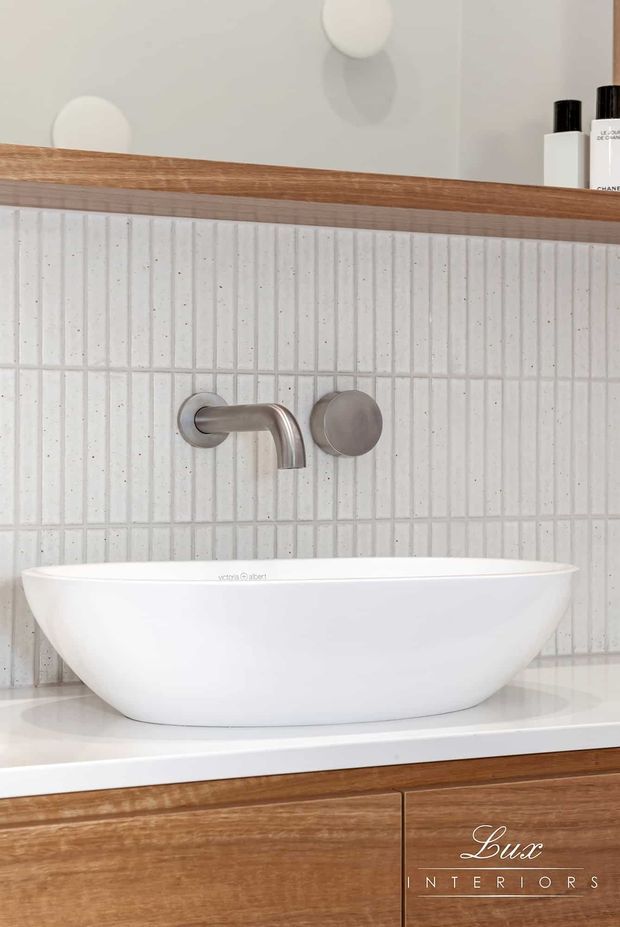 Close-up of a refined bathroom vanity featuring a white vessel basin on a white countertop, vertically ribbed white wall tiles, a brushed metallic wall-mounted tap, and warm timber cabinetry with a timber ledge and mirror above. Minimal toiletries are visible on the upper right.