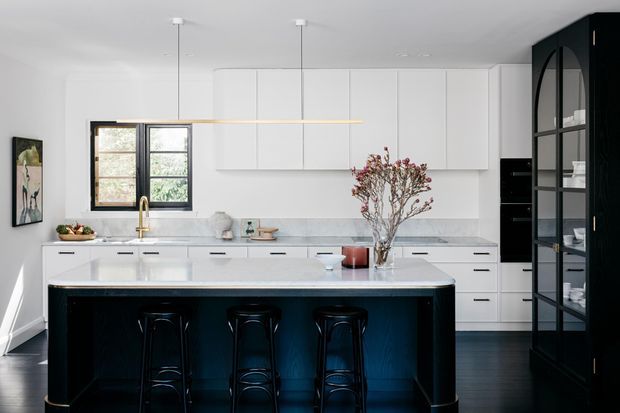 A bright contemporary kitchen with a large white stone island, three black stools, flat-panel white cabinetry, dark timber flooring, and a tall black built-in display cabinet with arched glass doors. A black-framed window, brass tapware, pendant lighting, and restrained floral styling add contrast.
