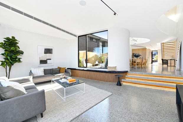Bright contemporary open-plan living room with a large grey sectional sofa, glass coffee table and pale rug. A curved white column, timber-lined built-in seating, expansive glazing, polished terrazzo-like flooring and illuminated steps lead toward a dining area and slatted timber screen. The space has a clean, airy and refined appearance.