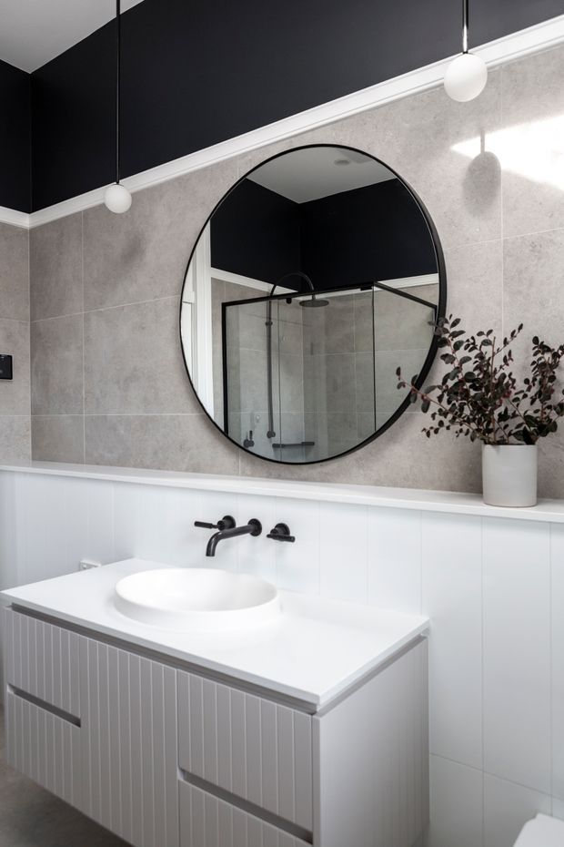 Contemporary bathroom vanity with a white vessel basin, wall-mounted matte black tapware, vertically fluted white cabinetry, and a white benchtop. The walls combine large light-grey stone-look tiles with white lower wall panelling and crisp white trim. A large round black-framed mirror reflects a glass shower enclosure, while simple spherical pendant lights and a potted branch add restrained decorative detail.