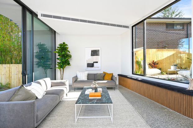 Bright contemporary living room with a large grey sectional sofa, matching loveseat, marble-look coffee table, indoor plants, and minimalist wall art. Floor-to-ceiling glazing on two sides opens views to a timber-fenced courtyard and neighbouring brick wall. A long built-in window seat or ledge with vertical timber slat cladding runs along the right side, and a linear ceiling air vent adds to the clean modern fit-out.