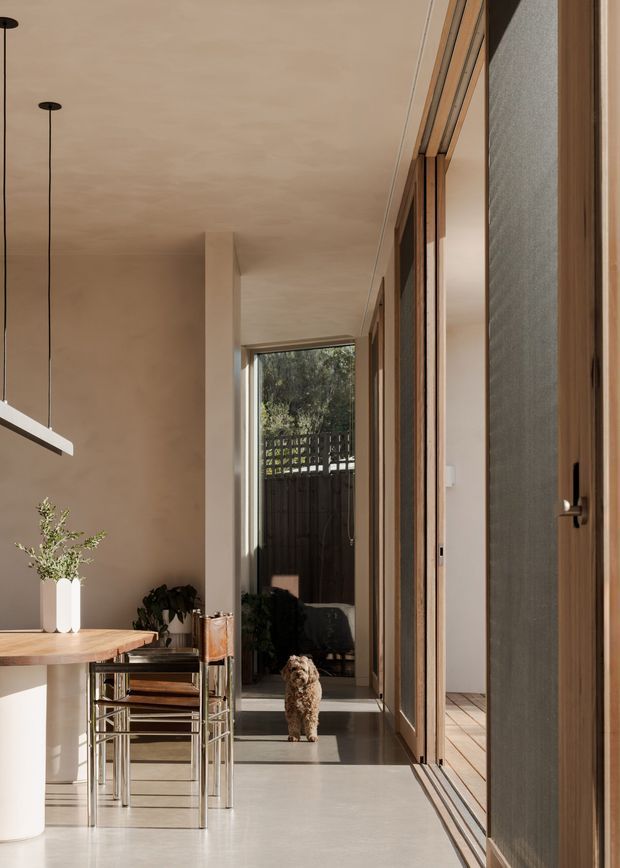 A warm, minimalist interior corridor adjoining a dining area. The space features a long timber dining table, slim metal-framed chairs, a linear pendant light, smooth beige plaster walls and ceiling, polished concrete flooring, and extensive timber-framed glazing and doors along the right side. Sunlight leads toward a small courtyard or outdoor area visible at the end of the passage.