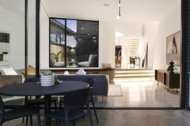 A bright contemporary open-plan living and dining area viewed from an adjoining space. The foreground features a dark round dining table with woven dining chairs, while the living area includes a dark sofa, rug, low timber-toned media cabinet, and potted greenery. Large black-framed glazing, polished concrete flooring, white walls, and a stepped transition lead toward a raised rear area with additional seating and a sculptural curved feature wall.