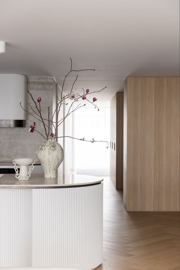A refined contemporary kitchen interior with a curved island featuring vertically fluted white cabinetry and a pale stone benchtop. The kitchen includes a textured stone backsplash, integrated stainless-steel rangehood, tall light-oak cabinetry, and warm herringbone timber flooring. A sculptural ceramic vase with branching flowers sits on the island, while soft daylight filters through sheer curtains in the background.
