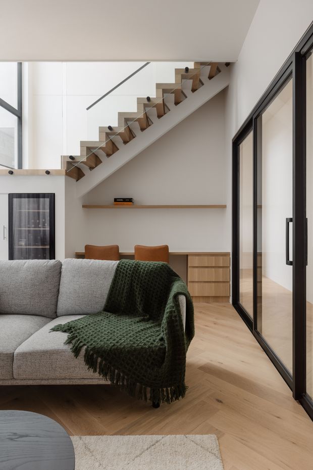 Contemporary living room with a grey sectional sofa, dark green knitted throw, light timber herringbone flooring, and a compact timber workspace or storage unit beneath a floating shelf. A sculptural timber staircase with clear glass balustrade rises through the space, while black-framed internal glazing lines the right side. The room is bright, refined, and warm with a neutral palette and natural timber accents.