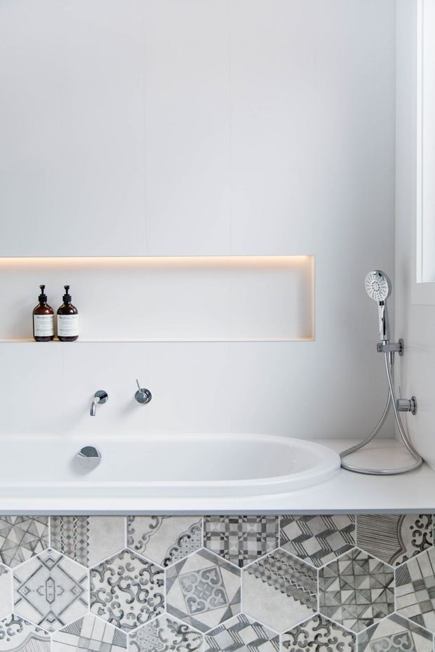 Bright, minimalist contemporary bathroom featuring a white built-in bathtub with wall-mounted chrome fixtures and a handheld shower. A recessed wall niche with warm integrated lighting displays two bottles, while the lower wall is finished with a striking patchwork of monochrome patterned tiles.