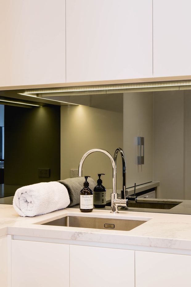 Close-up of a refined bathroom vanity with a white stone benchtop, integrated rectangular basin, chrome gooseneck tapware, white flat-panel cabinetry, and a large mirrored wall cabinet with concealed linear lighting. Neutral grey-beige wall finishes and warm under-cabinet illumination create a polished, contemporary atmosphere.