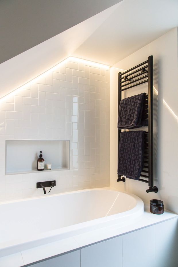 Bright contemporary bathroom with a built-in white bathtub beneath a sloped ceiling. White glossy brick-pattern wall tiles surround a recessed shower or bath niche, with warm concealed LED lighting along the ceiling line. A black wall-mounted heated towel rail with dark towels provides contrast, while white built-in cabinetry forms the bath surround.