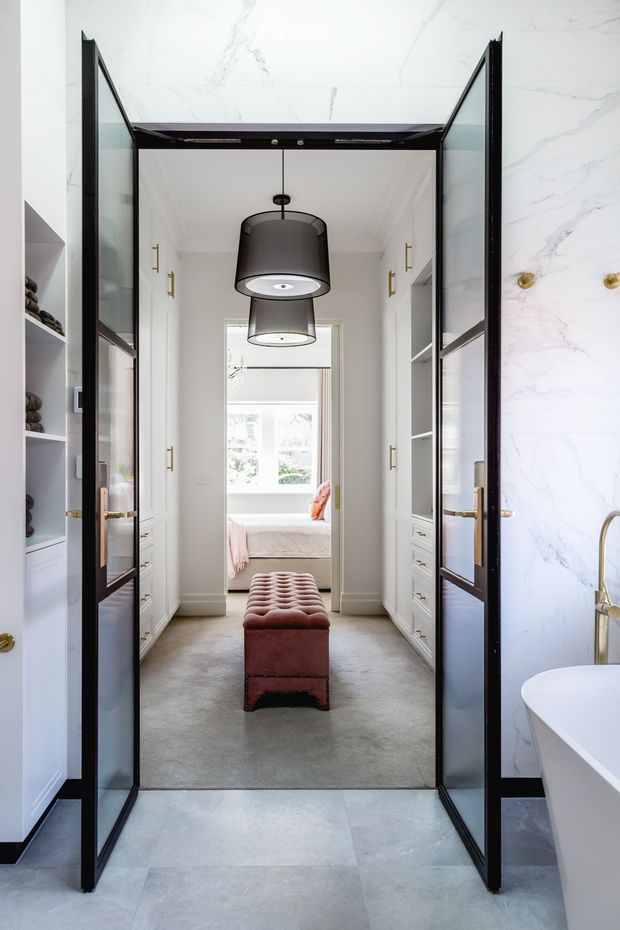 A bright, refined bathroom foreground opens through black-framed glass doors into a carpeted dressing corridor and bedroom beyond. The bathroom features large white marble-look wall surfaces, pale stone flooring, white built-in cabinetry, brass hardware and a freestanding white bathtub. A dark pendant light hangs over a tufted burgundy leather bench in the dressing area.