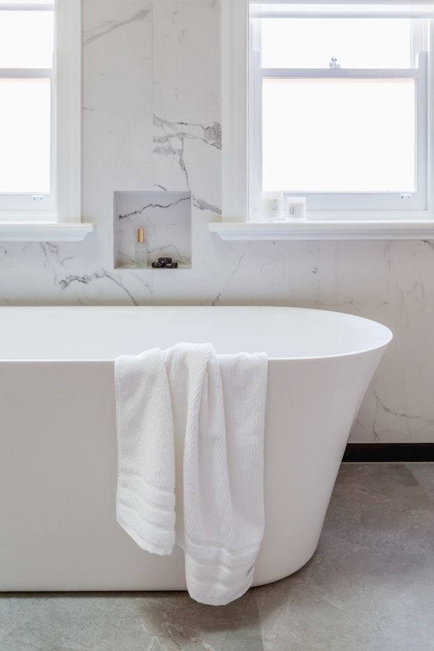 A bright, refined bathroom featuring a freestanding white oval bathtub, white marble-look wall surfaces with grey veining, two white-framed windows, a recessed wall niche with toiletries, and a grey stone-look floor. White towels hang over the bath.