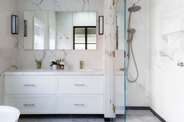 Bright, refined bathroom featuring a double vanity with white shaker-style cabinetry, white marble-look wall surfaces, brass tapware and hardware, a large mirrored cabinet, and a glass-enclosed walk-in shower with a brushed metal shower set. Black trim accents and grey stone-look flooring add contrast.