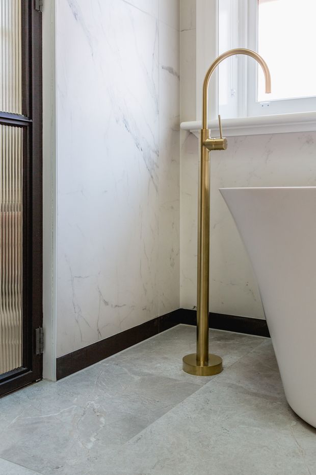 Close-up of a refined bathroom corner featuring a freestanding white bathtub, a tall brushed brass floor-mounted bath filler, marble-look wall cladding, grey veined stone-look flooring, dark skirting, and a large white-trimmed window. A dark-framed textured glass door is visible at the left.