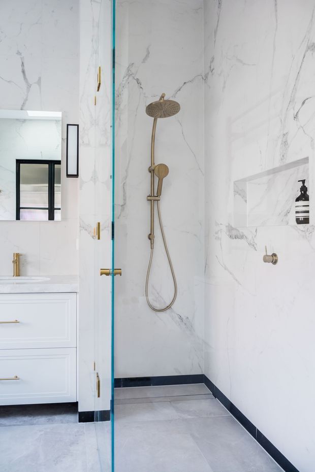 Bright contemporary bathroom featuring a frameless glass walk-in shower with brushed brass shower fittings, large white marble-look wall tiles with grey veining, pale grey floor tiles, dark skirting accents, and a white vanity with brass hardware. A mirrored cabinet and recessed shower niche are visible.