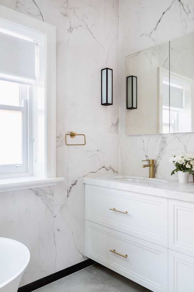 Bright, refined bathroom featuring full-height white marble-look wall surfaces with grey veining, a white paneled vanity with gold hardware, integrated white basin, brass tapware, frameless mirror, and slim black wall sconces. A window with a white roller blind provides natural light, while a freestanding white bathtub is partially visible.