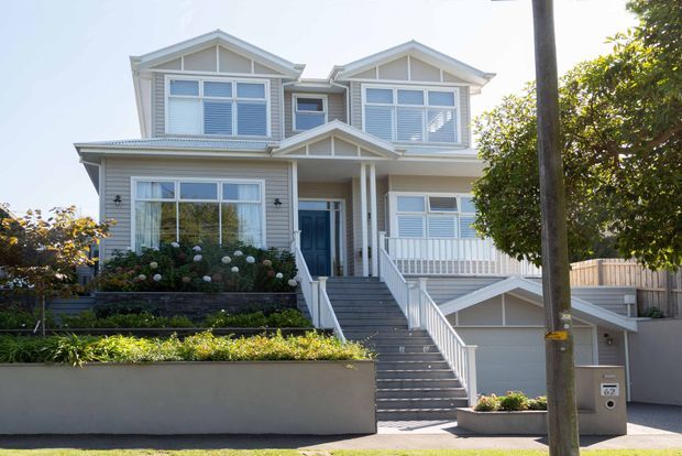 Front exterior view of a large two-storey light-coloured home with prominent front gables, covered entry porch, broad staircase, white-framed windows, decorative balustrades, and an integrated garage. The landscaped front garden and boundary wall frame the facade.