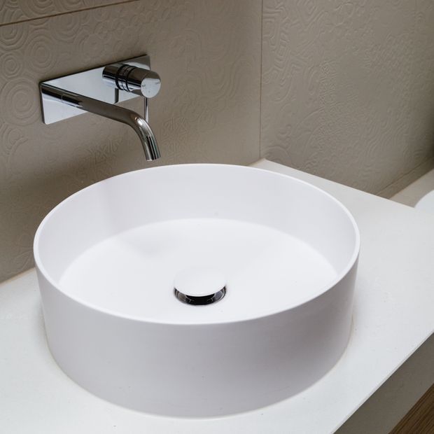 Close-up of a contemporary bathroom vanity featuring a round white vessel basin on a white benchtop, paired with a wall-mounted chrome tap and drain. The wall has a subtle embossed textured finish.