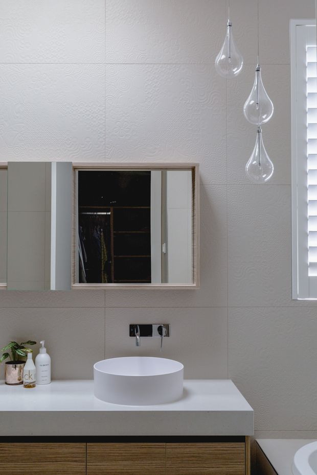 A refined contemporary bathroom vanity featuring a white vessel basin on a pale stone benchtop, a wood-grain floating cabinet, textured white wall tiles, a mirrored cabinet, a wall-mounted chrome tap, pendant lights with clear teardrop shades, and white plantation shutters. A partial bathtub is visible at the lower right.
