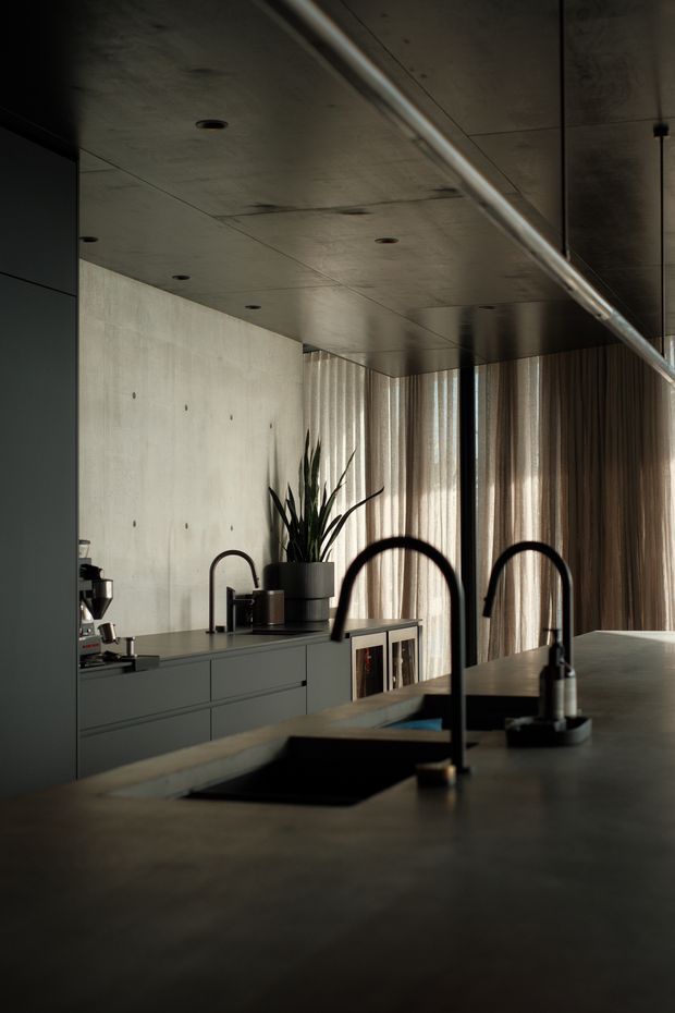 A moody contemporary kitchen interior viewed across a large island. The space features dark grey fixed cabinetry, a broad dark benchtop with integrated sinks and tall black gooseneck taps, a concrete-look wall and ceiling, timber-toned vertical curtains, and restrained recessed lighting. A coffee machine and potted plant sit on the rear counter, with warm daylight filtering through the curtains.