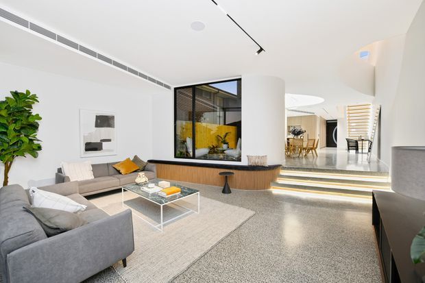 Bright contemporary open-plan living room with two grey sofas, a light rug and glass coffee table, facing a curved timber-clad built-in feature wall and large internal window. Steps lead to an elevated dining and circulation area, with white walls, polished speckled flooring, linear track lighting and abundant natural light.