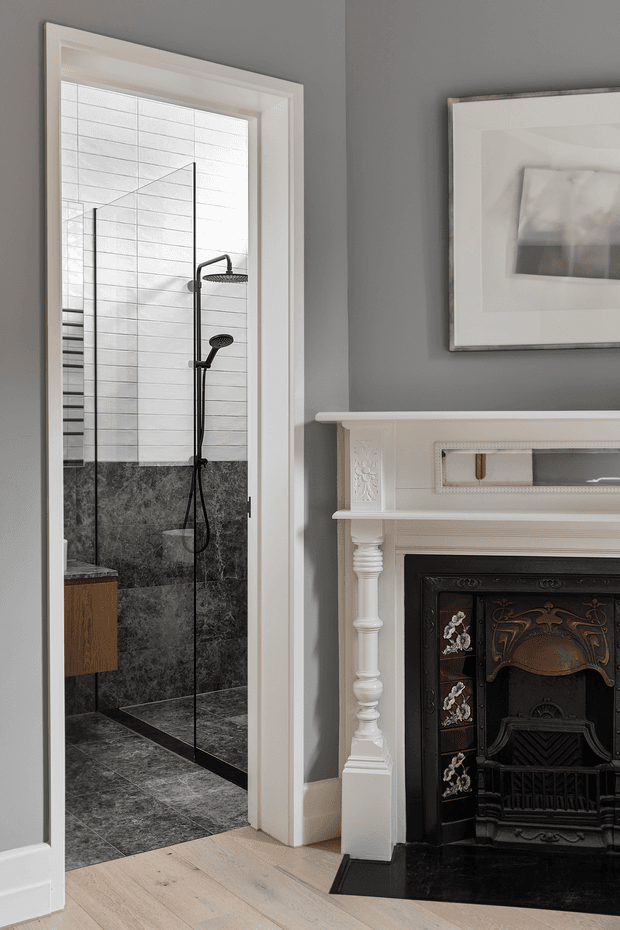 A refined interior detail showing a walk-in shower viewed through an open doorway beside an ornate traditional fireplace. The shower has a frameless glass screen, black fixtures, white rectangular wall tiles, dark stone-look wall and floor tiles, and a floating timber vanity partially visible at left. The surrounding room features grey walls, pale timber flooring, white decorative fireplace joinery, and a black cast-iron firebox with floral detailing.