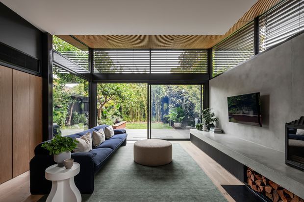 A contemporary living room with a deep navy sectional sofa, round upholstered ottoman, pale sculptural side table, and a long built-in concrete media console with fireplace and timber log storage. Full-height black-framed glazing and horizontal louvres open the room to a leafy garden, while a timber slatted ceiling and timber wall panelling add warmth. The space has a refined, calm indoor-outdoor character.