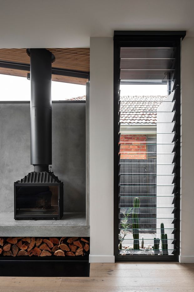 Interior view of a contemporary living space featuring a black freestanding wood-burning fireplace with a tall flue, set into a grey concrete hearth and wall surround. Firewood is stored beneath the hearth. A tall black-framed louvred window or glazed door provides ventilation and views to an adjoining brick building and courtyard planting. Timber slatted shading is visible above the left-side glazing, and pale timber flooring runs throughout.