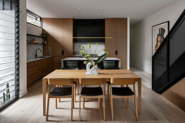 A warm, contemporary open-plan kitchen and dining area viewed from the living space. The kitchen features full-height natural timber cabinetry, a dark recessed rangehood and splashback zone, pale stone benchtops, open shelving, and a compact island with seating. In the foreground, a timber dining table with matching timber chairs and dark seats sits beneath simple pendant lights. Light timber flooring, strong daylight, and a black mesh staircase balustrade complete the refined interior.
