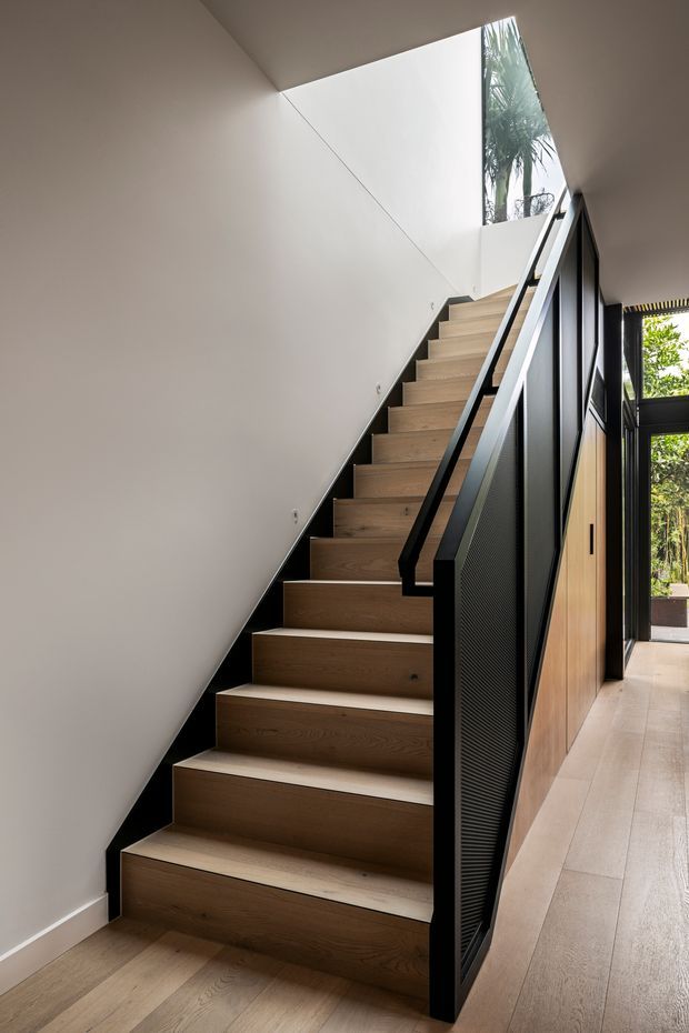 Interior view of a contemporary stairway with broad light timber treads and matching timber flooring. The staircase has a black metal balustrade and perforated screen, adjacent warm timber panelled joinery, smooth off-white walls, and strong natural light entering from tall glazing and an upper opening.