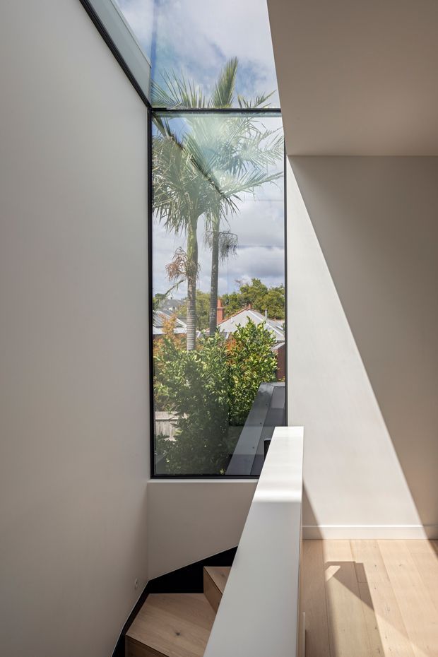 A bright modern stairway with pale timber treads, crisp white walls, and a white balustrade. A tall, narrow black-framed window brings in strong sunlight and views of palm trees and neighbouring rooftops, creating a vertical connection to the outdoors.