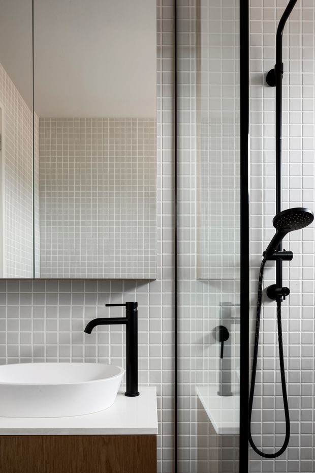 A compact contemporary bathroom featuring a white vessel basin on a timber vanity, matte black wall-mounted tapware, and a black-framed glass shower enclosure with handheld and overhead shower fittings. Small pale grey square tiles cover the walls, creating a clean, restrained finish.