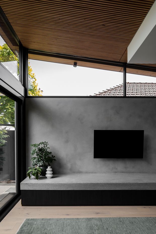 Contemporary living room media wall with a long built-in concrete bench and dark cabinetry below, a wall-mounted television, black-framed floor-to-ceiling glazing, and a warm slatted timber ceiling. Indoor planting and a muted rug soften the predominantly charcoal, timber, and grey palette. A tiled roof and greenery are visible outside through the glazing.