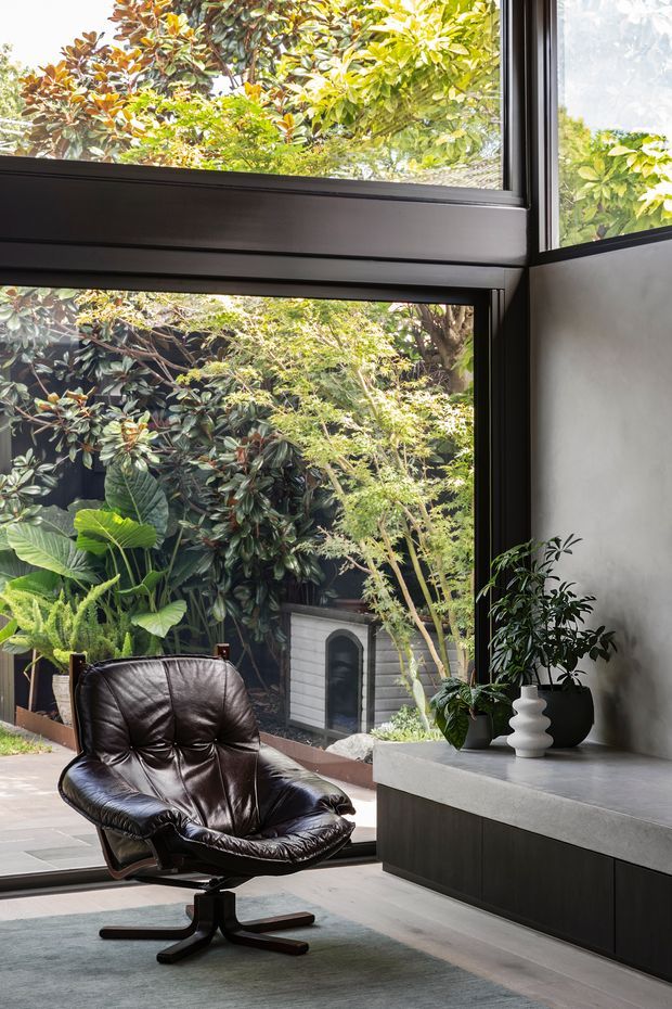 A refined contemporary interior corner with a black leather lounge chair beside floor-to-ceiling black-framed glazing. The glazing overlooks a dense landscaped garden, while a minimalist dark built-in cabinet and pale stone or concrete benchtop run along the wall. Potted plants add greenery, and the room has a calm, sophisticated palette of charcoal, grey, and natural green.