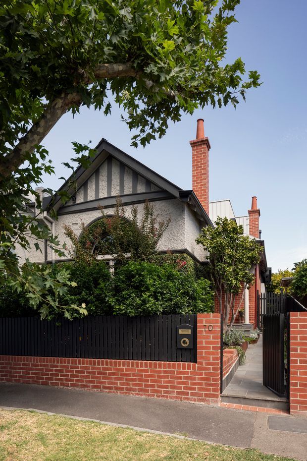 Street-facing exterior view of a traditional suburban residence with a steep gabled roof, decorative half-timbered gable detailing, textured rendered walls, red brick chimneys, and a landscaped front garden behind a black vertical-slat fence. A brick pillar and pedestrian gate open onto a narrow paved entry path.