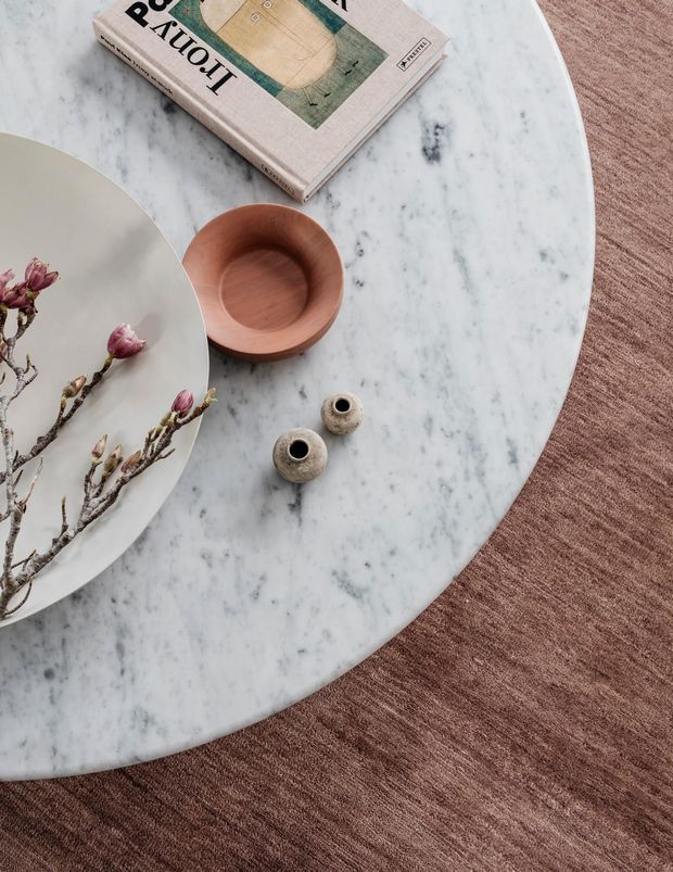 Close-up styling vignette of a round white-veined marble tabletop over a warm brown textured rug. The table holds a pale ceramic plate with flowering branches, a small carved wooden bowl, two small textured ceramic vessels, and a design book, creating a refined, organic tabletop arrangement.